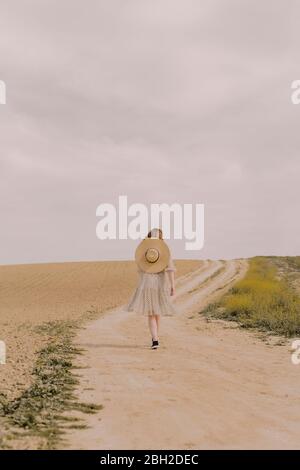 Frau mit Strohhut und Vintage-Kleid zu Fuß auf einer abgelegenen Feldstraße in der Landschaft Stockfoto