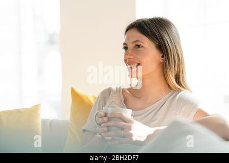 Glückliche junge Frau, die sich auf der Couch entspannt und vor dem Fenster einen Kaffeebecher hält Stockfoto