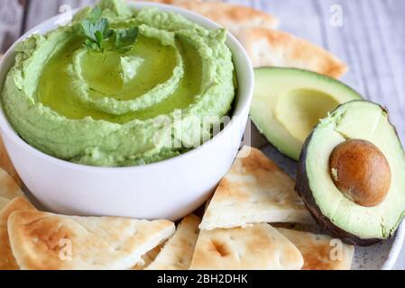 Veganer Avocado Hummus, hergestellt aus Kichererbsen, Avocados und Tahini, mit Olivenöl. Mit Petersilie garniert und mit Pita-Brot auf einem rustikalen Tisch serviert. Stockfoto