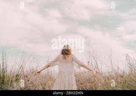 Rückansicht der Frau im Vintage-Kleid mit ausgestreckten Armen auf einem abgelegenen Feld auf dem Land Stockfoto