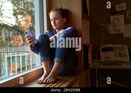 Mädchen, das barfuß auf der Fensterbank am Abend sitzt und Smartphone anschaut Stockfoto