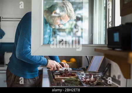 Reifer Mann, der Salat am offenen Fenster in seiner Küche zubereitet Stockfoto