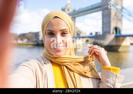 Porträt einer lächelnden jungen Frau, die vor der Tower Bridge ein Selfie macht, London, Großbritannien Stockfoto