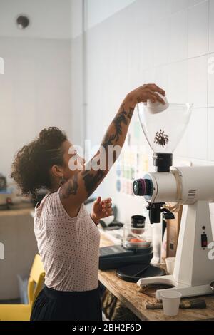 Frau, die in einer Kaffeerösterei arbeitet und Kaffeebohnen in eine Kaffeemaschine gießt Stockfoto