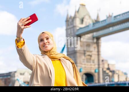 Porträt einer lächelnden jungen Frau, die mit dem Smartphone vor der Tower Bridge, London, UK, Selfie macht Stockfoto