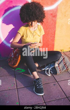 Junge Frau mit Afro-Frisur sitzt an der Graffiti-Wand und mit dem Smartphone Stockfoto