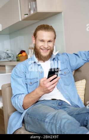 Glücklicher junger Mann, der sich auf dem Sofa mit dem Smartphone entspannt und zu Hause die Kamera anschaut. Stockfoto