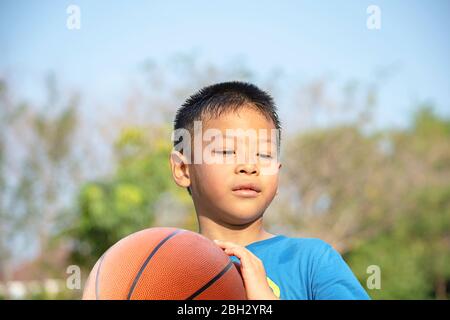 Asian Boy Holding ein Basketball Ball Hintergrund verschwommen Bäume. Stockfoto