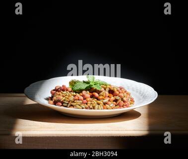 Bohnen und Roggen mit Minzblättern auf einem weißen Teller, auf einem Holzhackbrett, beleuchtet durch direktes Sonnenlicht Stockfoto