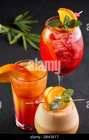 Drei alkoholische Cocktails, rot, orange und gelb auf schwarzem Hintergrund. Verschiedene alkoholische Getränke mit Orangenscheiben Stockfoto