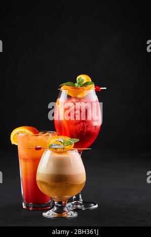 Drei alkoholische Cocktails, rot, orange und gelb auf schwarzem Hintergrund. Verschiedene alkoholische Getränke mit Orangenscheiben. Kopierbereich Stockfoto