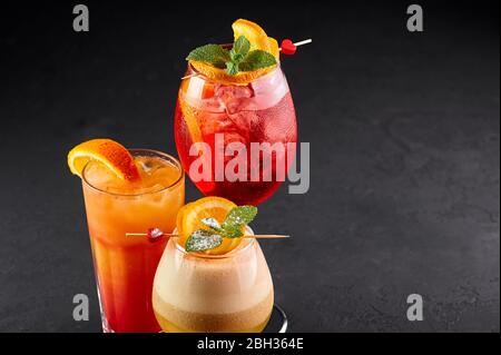 Drei alkoholische Cocktails, rot, orange und gelb auf schwarzem Hintergrund. Verschiedene alkoholische Getränke mit Orangenscheiben. Kopierbereich Stockfoto