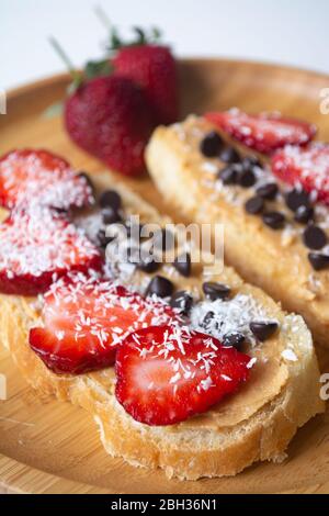 Erdnussbutter Aufstrich mit Schokoladenstückchen, Erdbeeren, Kokosflocken und hausgemachtem Brot auf einem Holzteller Stockfoto