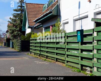 Grün gebeizt Holzzaun. Horizontale Planken. Vertikale Holzpfosten. Weiße Stuckhaus Außenfassade. Roll-down-Fensterläden. Grüne Hecke & Kiefer Stockfoto