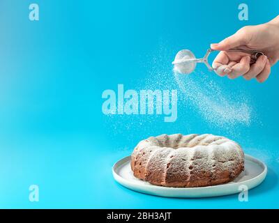 Frau Hand Streuseln Puderzucker über frisch hausgemachte Kuchen gebündelt. Puderzucker fällt auf frischen perfekten Bunt-Kuchen auf blauem Hintergrund. Platz für Text kopieren. Ideen und Rezepte für Frühstück oder Dessert Stockfoto