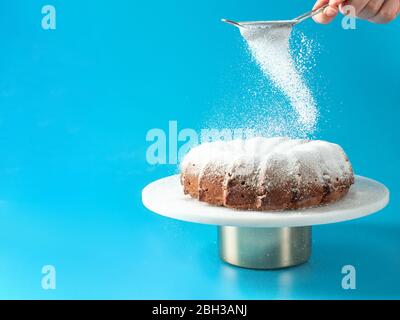 Frau Hand Streuseln Puderzucker über frisch hausgemachte Kuchen gebündelt. Puderzucker fällt auf frischen perfekten Bunt-Kuchen auf blauem Hintergrund. Platz für Text kopieren. Ideen und Rezepte für Frühstück oder Dessert Stockfoto