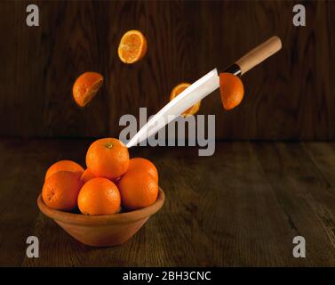 Messer schneiden Orangen. Viele Orangen auf der Schale - dunkler Hintergrund. Stillleben Früchte im Vintage-Stil. Fliegende Orangen auf der Schale dunkel Foto Stockfoto