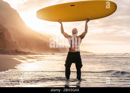 Happy Fit Senior mit Spaß beim Surfen bei Sonnenuntergang - sportliches bärtiger Mann Training mit Surfbrett am Strand Stockfoto