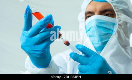 Nahaufnahme des latein-Arztes in Bioprotective-Anzug, Gesichtsmaske und blauen Handschuhen, die eine Blutprobe in einem Reagenzglas mit einer Spritze auf weißem Hintergrund entnehmen Stockfoto