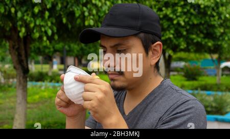 Nahaufnahme des lateinischen Mannes auf der Straße starrend auf eine weiße Gesichtsmaske, die er mit beiden Händen hält, um sie anzuziehen Stockfoto