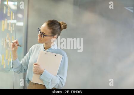 Junge Geschäftsfrau in Brillen, die neben der Glaswand steht und ihre Pläne im Büro auf Haftnotizen schreibt Stockfoto