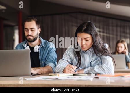 Foto von multinationalen ernsthaften Studenten, die Laptop und das Schreiben in Übungsheft während des Studiums im Klassenzimmer benutzen Stockfoto