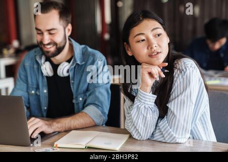 Foto von multinationalen jungen Studenten, die Laptop und Schreiben in Übungsheft während des Studiums im Klassenzimmer Stockfoto