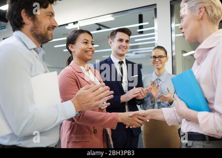 Zwei Geschäftsfrauen, die nach erfolgreicher Vereinbarung mit anderen Geschäftsleuten, die stehen und klatschen beim Büro die Hände schütteln Stockfoto