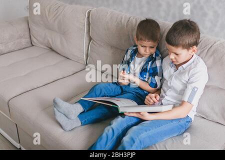 Zwei Brüder lesen ein Buch, während sie während der Quarantäne auf einem Sofa im Wohnzimmer sitzen. Das Konzept der sozialen Distanz, Selbstisolierung und Heimat scho Stockfoto