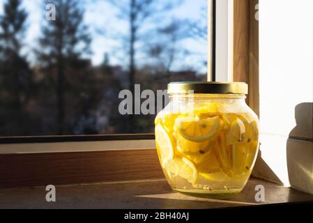 Limonade mit Scheiben Zitronen in einem großen Glas auf der Küchenfensterbank in der Morgensonne. Glas auf der Fensterbank mit hausgemachter Limonade. Gesund Stockfoto