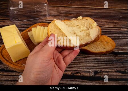 Jemand breitet ein wenig Butter mit einem Messer auf frischen Weizenstoast, ein Stück Butter in einer hölzernen Butterdose und frischen Weizen Toast auf einem hölzernen Backgr aus Stockfoto