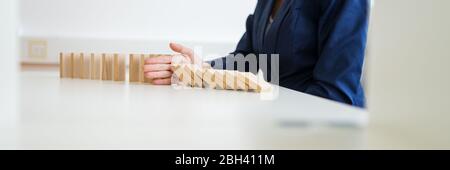 Weitblick-Bild einer nicht erkennbaren Geschäftsfrau, die an ihrem weißen Büroschreibtisch sitzt und mit ihrer Hand zusammenbrechende Dominos in einem konzeptuellen Bild von c Stockfoto