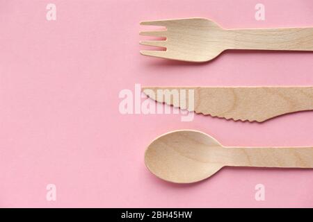 Öko Besteck aus Kiefernholz. Einweggeschirr aus natürlichen Materialien. Holzlöffel, Gabel und Messer gegen Rosa mit Kopierraum. Vertikales Bild. Stockfoto