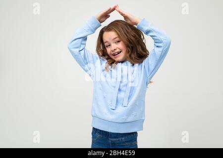 Lächelndes Teenager-Mädchen in lässigem blauen Hoodie legte ihre Hände über ihren Kopf in Form eines Daches auf einem weißen Hintergrund mit Kopierraum. Stockfoto