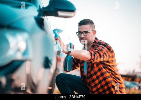 Porträt eines jungen Mannes, der sein Auto mit einem Tuch und Spray in einer Flasche polieren, im Freien. Stockfoto