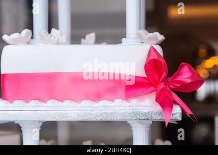 Nahaufnahme von mehrstufigen weißen Fondant Sahnetorte mit rosa Band Dekoration auf Standansicht durch Fenster der Bäckerei Stockfoto