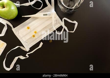 Weiße medizinische Gegenstände in einer Ecke auf einem dunklen Hintergrund, eine schützende medizinische Maske mit Krawatten, Pillen, ein Thermometer, ein Glas Wasser, einen Apfel Stockfoto