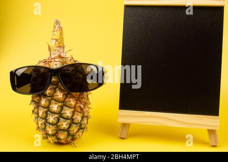 Ananas trägt Sonnenbrille mit leerem schwarzen Brett auf gelbem Hintergrund Stockfoto