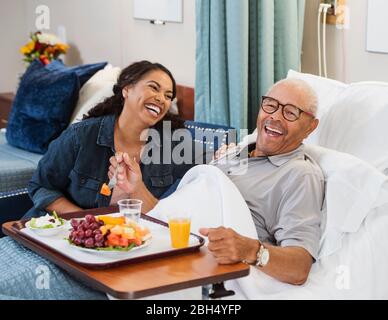 Lachende Frau, die neben einem älteren Mann sitzt und im Bett Obst isst Stockfoto