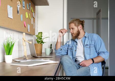Konzentrierter junger Mann, der auf den Laptop-Bildschirm schaut, der am Schreibtisch des Heimbüros sitzt. Stockfoto