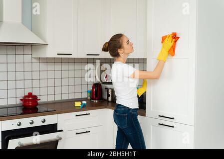 Das Konzept der Frühjahrsputz, Reinigungsfirma. Junge Frau Reinigung einer Oberfläche von weißen Küchenwand Schrank, tragen Gummi-Schutzhandschuhe gelb, mit Lappen. Haus, Hauswirtschaftskonzept. Stockfoto