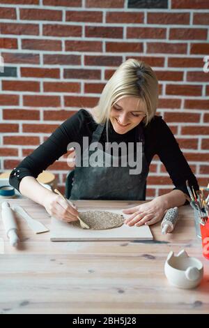 Glücklich lächelnde Frau Herstellung Keramikplatte, tippen Sie mit Spatel. Konzept für Frau in freiberuflicher, geschäftlicher. Handwerkliches Produkt. Verdienen Sie mehr Geld, Side Hustle Stockfoto