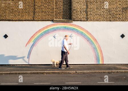 Camberwell, London, Großbritannien. April 2020. Ein Mann geht am frühen Morgen mit seinem Hund an einem Wandbild eines Regenbogens vorbei. Seit der Pandemie Covid-19 ist der Regenbogen zu einem Symbol der Unterstützung für das NHS-Personal und alle Pflegekräfte in Großbritannien geworden. Dieser Regenbogen wird von Louis Young gemalt, um dem NHS-Personal zu danken, das sich um seinen Vater kümmerte und vor kurzem aus dem Krankenhaus kam. Kredit: Tom Leighton/Alamy Live News Stockfoto