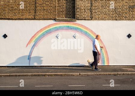 Camberwell, London, Großbritannien. April 2020. Ein Mann geht am frühen Morgen mit seinem Hund an einem Wandbild eines Regenbogens vorbei. Seit der Pandemie Covid-19 ist der Regenbogen zu einem Symbol der Unterstützung für das NHS-Personal und alle Pflegekräfte in Großbritannien geworden. Dieser Regenbogen wird von Louis Young gemalt, um dem NHS-Personal zu danken, das sich um seinen Vater kümmerte und vor kurzem aus dem Krankenhaus kam. Kredit: Tom Leighton/Alamy Live News Stockfoto