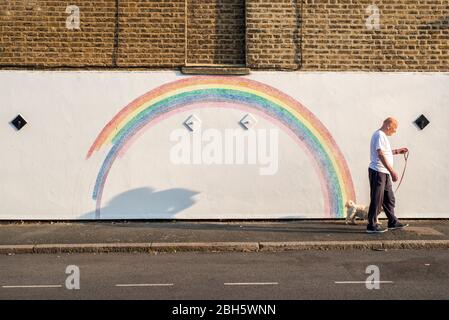 Camberwell, London, Großbritannien. April 2020. Ein Mann geht am frühen Morgen mit seinem Hund an einem Wandbild eines Regenbogens vorbei. Seit der Pandemie Covid-19 ist der Regenbogen zu einem Symbol der Unterstützung für das NHS-Personal und alle Pflegekräfte in Großbritannien geworden. Dieser Regenbogen wird von Louis Young gemalt, um dem NHS-Personal zu danken, das sich um seinen Vater kümmerte und vor kurzem aus dem Krankenhaus kam. Kredit: Tom Leighton/Alamy Live News Stockfoto