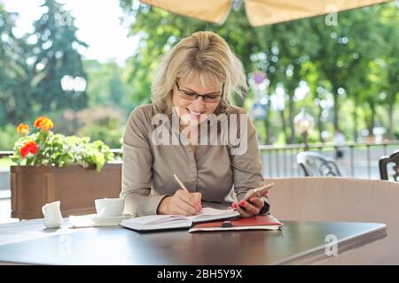 Business schöne Frau Schreiben in Business-Notebook mit Smartphone, lächelnde Frau sitzen im Sommer im Freien Café, Kaffeepause Stockfoto