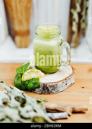 Gesunder Vitaminshake mit Spinat, Ingwer und Mandelmilch in einem stilvollen Glas, verziert mit Zitrone und Spinatblatt auf einem Holzteller, grün, gelb Stockfoto