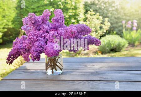 Schöner Blumenstrauß von lila lila Blüten in einem Glas auf einem Holztisch im Garten Stockfoto