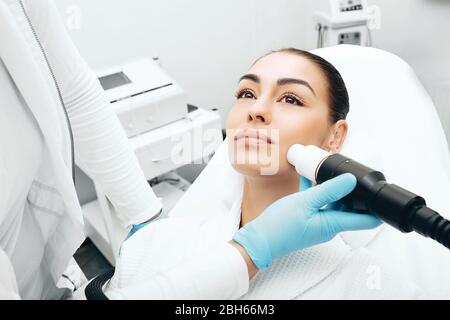 Kosmetikerin mit einer akustischen Wellenmaschine verjüngt die Haut des Gesichts einer Frau. Schmerzlose Hautverjüngung Stockfoto