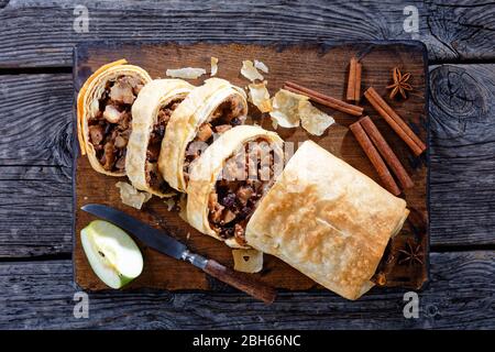 Deutscher Apfelstrudel mit karamellisierten Äpfeln, Nüssen, Zimt, Rosinen auf einem Holzbrett mit Sternanis serviert, Zimtstangen, Messer auf einem ru Stockfoto